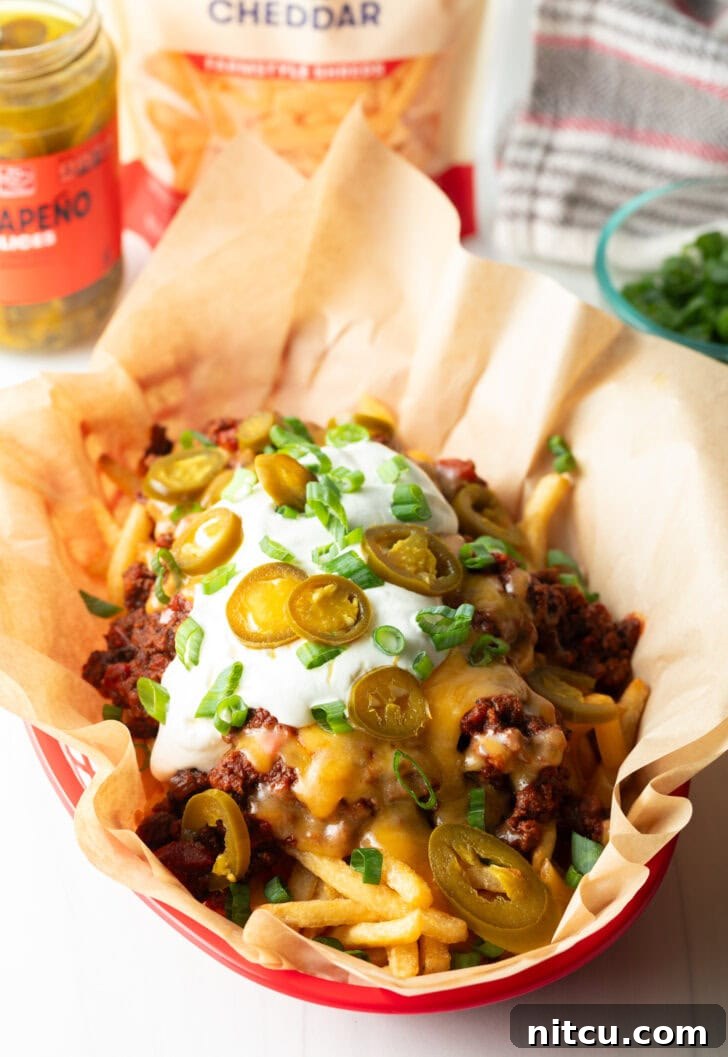Smothered Chili Cheese Fries 11 A rustic basket lined with parchment paper, bursting with crispy french fries lavishly topped with rich chili and melted cheddar cheese. It's elegantly garnished with creamy sour cream, vibrant chopped green onions, and fiery sliced jalapeños, showcasing a truly indulgent dish.