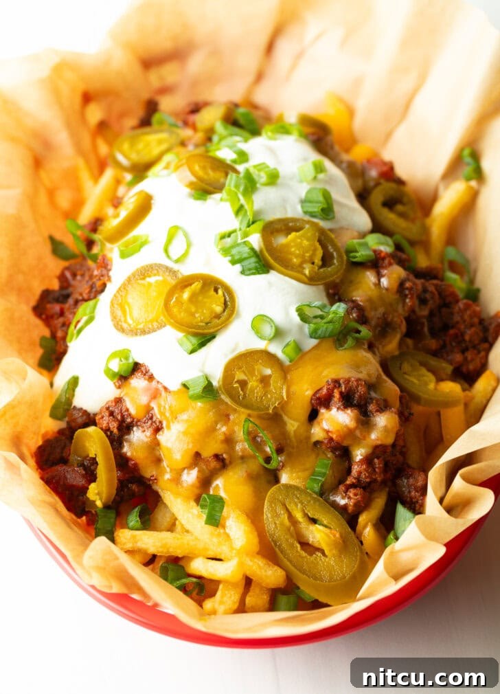 Smothered Chili Cheese Fries 12 A rustic basket lined with parchment paper, filled with crispy french fries generously loaded with rich chili, melted cheddar cheese, cool sour cream, fresh chopped green onions, and vibrant sliced jalapeños. The dish is presented beautifully, ready to be enjoyed.