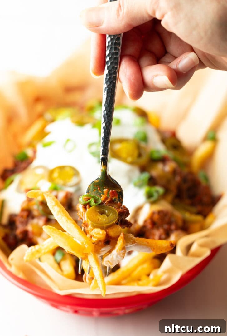 Smothered Chili Cheese Fries 13 A rustic basket lined with parchment paper, filled with crispy french fries lavishly topped with rich chili, perfectly melted cheddar cheese, cool sour cream, fresh chopped green onions, and vibrant sliced jalapeños. A hand holding a forkful, showcasing the delicious layers, is visible in the foreground.