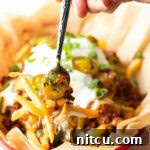 Basket with parchment paper, filled with crispy fries topped with chili and melted cheddar cheese, sour cream, chopped green onions, and jalapeno slices. A hand with forkful of fries is showing to camera.