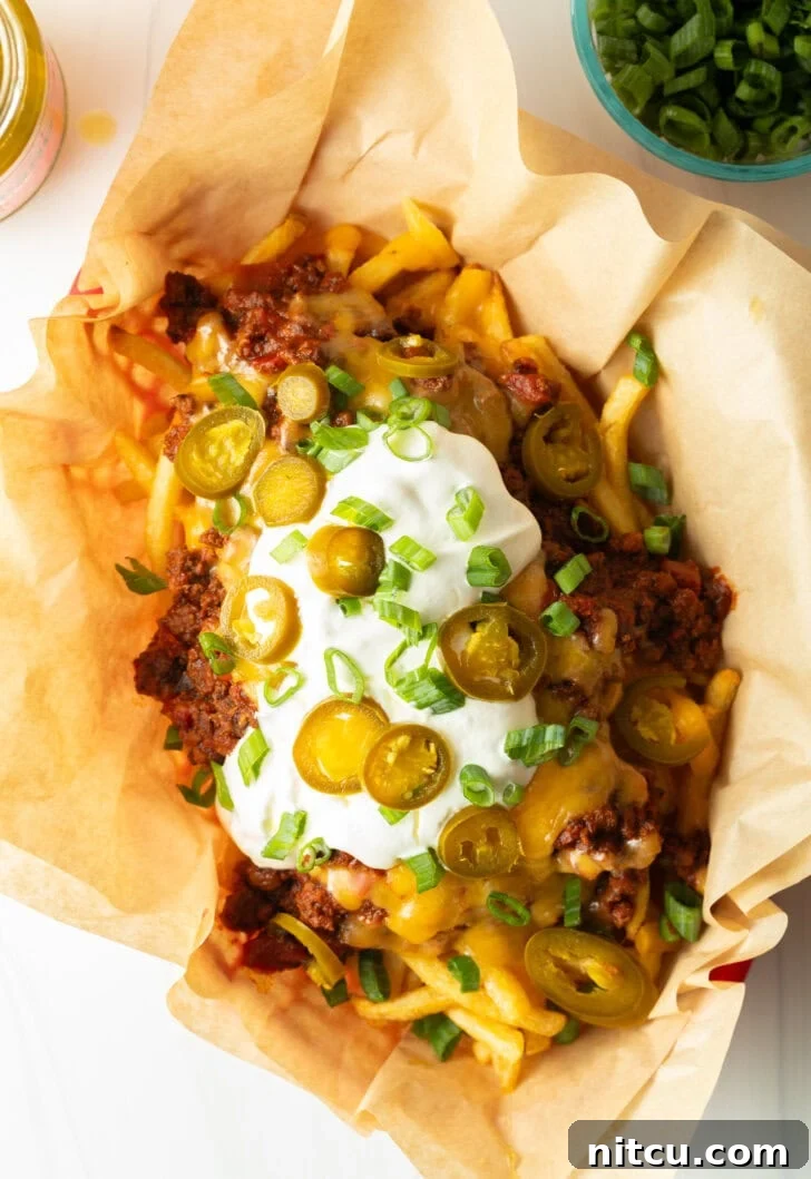 Smothered Chili Cheese Fries 3 A rustic basket lined with parchment paper, filled with crispy french fries generously covered with rich chili and a thick layer of melted cheddar cheese. It's garnished with cool sour cream, vibrant chopped green onions, and fiery sliced jalapeños, ready to be served.