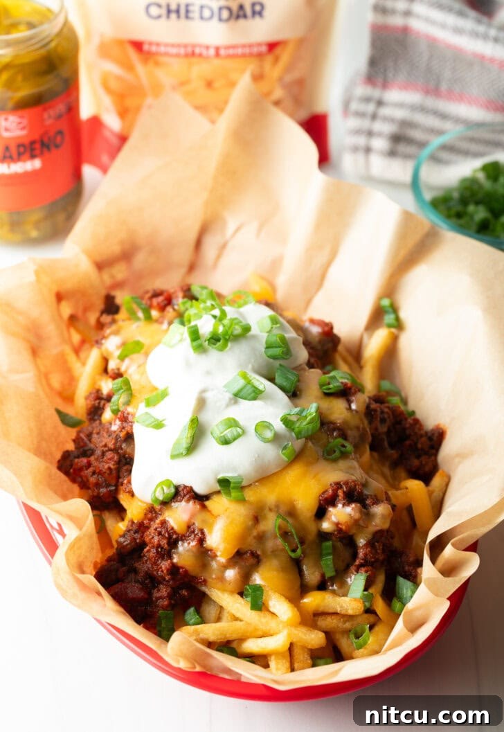 Smothered Chili Cheese Fries 10 A rustic basket lined with parchment paper, showcasing crispy french fries topped with hot chili, perfectly melted cheddar cheese, creamy dollops of sour cream, and a scattering of fresh chopped green onions.