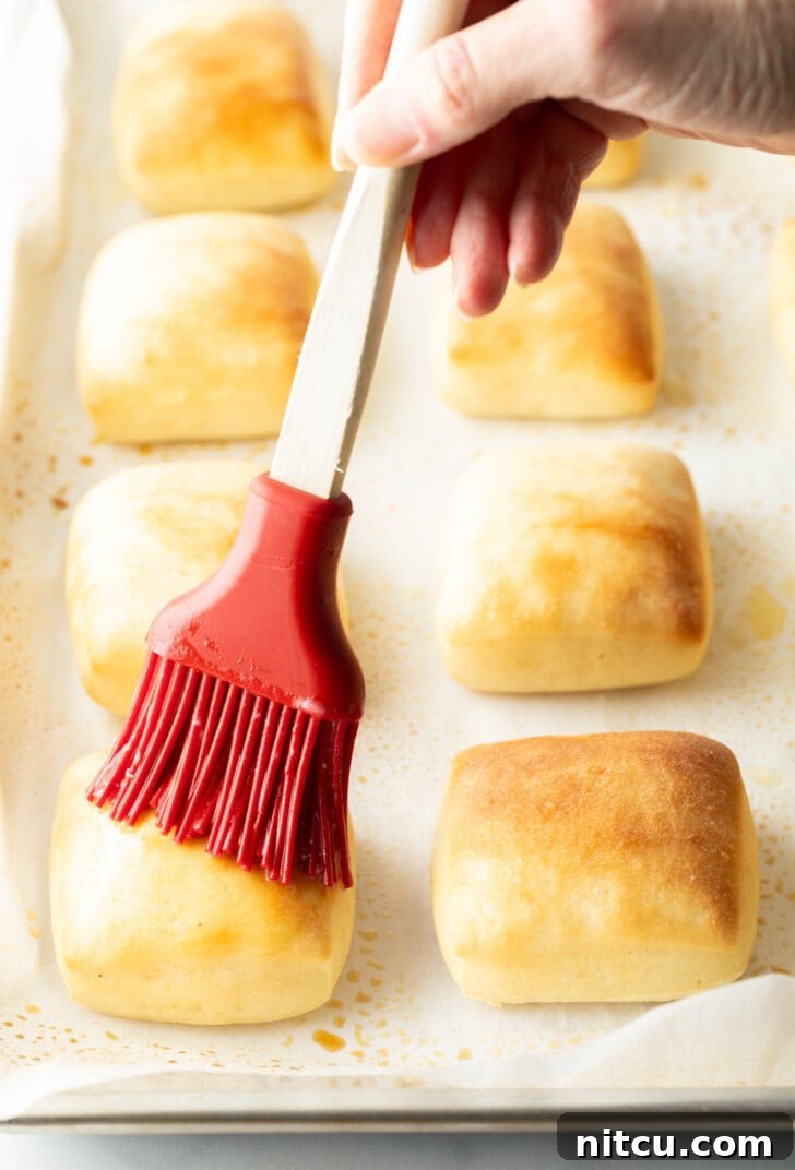 Hand with red basting brush, brushing melted butter over baked yeast rolls on a baking sheet.