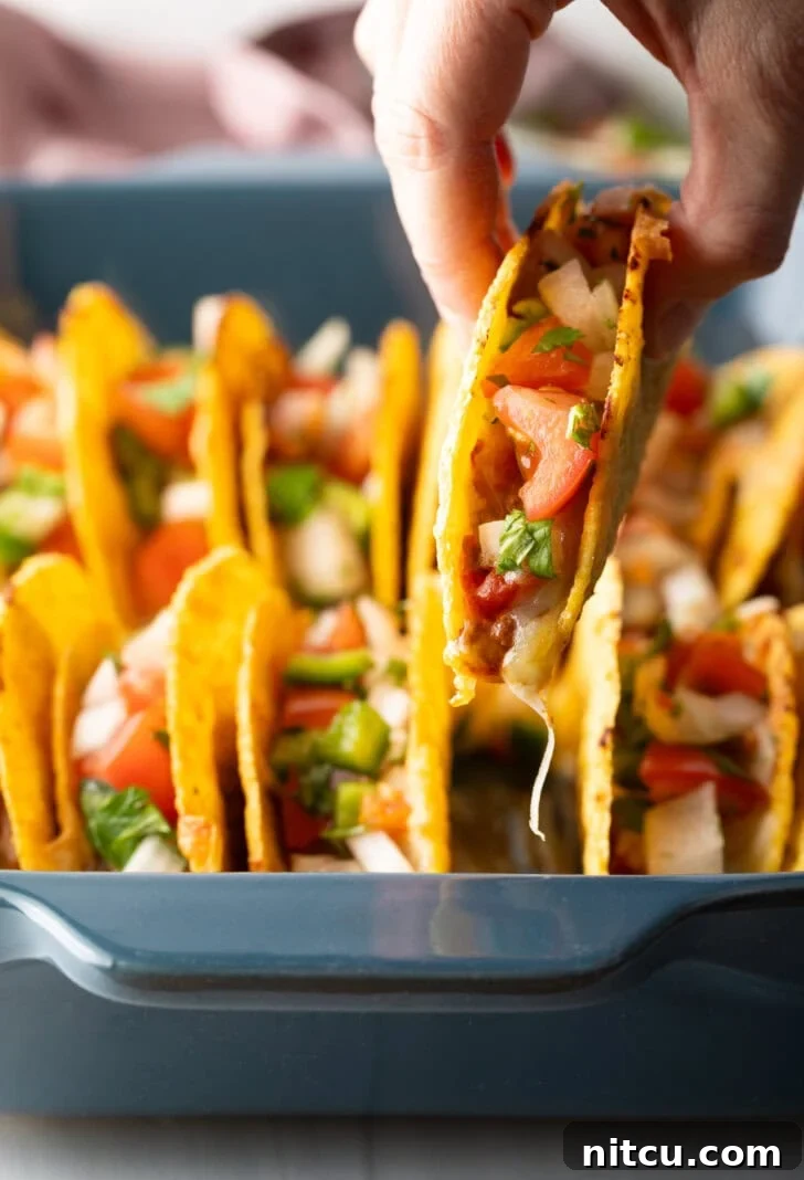Effortless Baked Tacos 3 A hand reaching into a baking dish to pick up one of the freshly baked, loaded tacos.