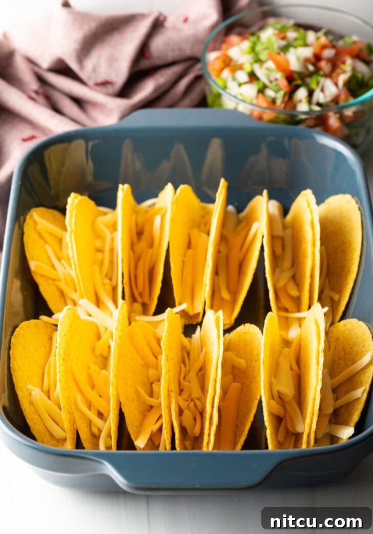Effortless Baked Tacos 6 Two rows of a dozen yellow corn hard shell tacos, standing upright in a large baking dish, with shredded cheese sprinkled in the bottoms.