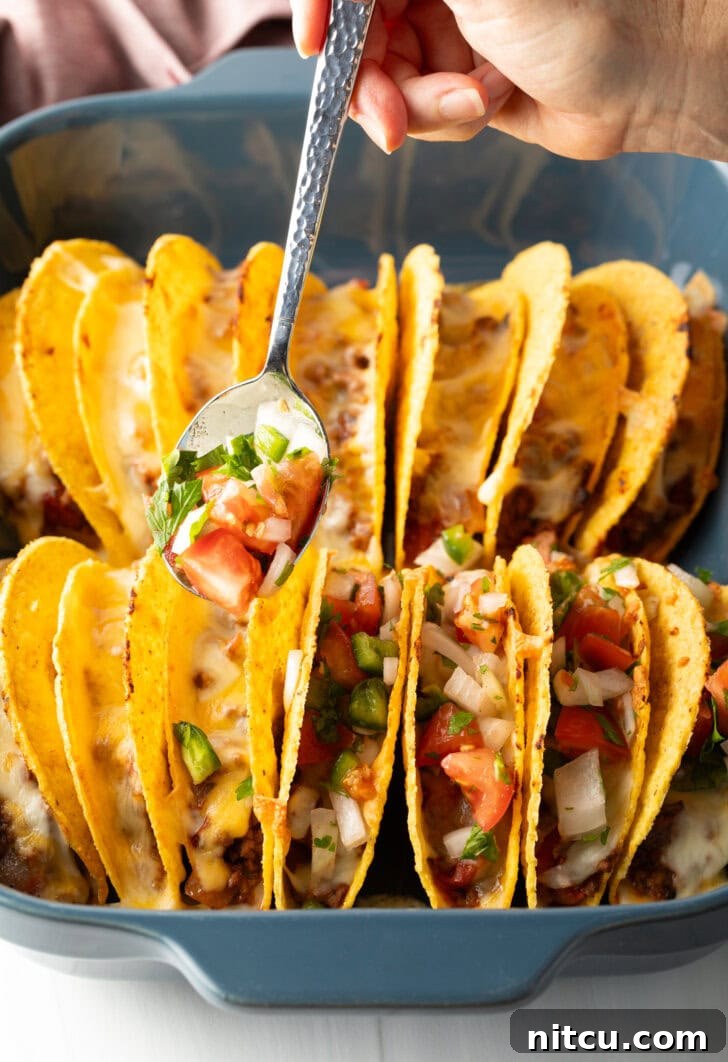Effortless Baked Tacos 9 A spoon adding fresh pico de gallo to a row of hard shell tacos, nestled in a baking dish.
