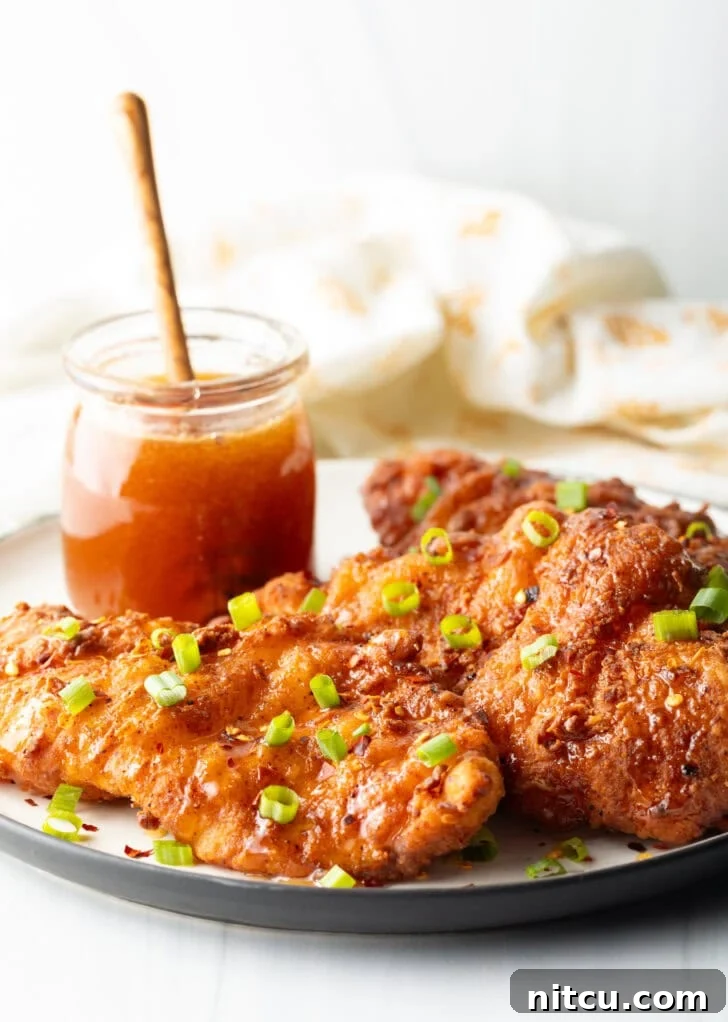 Sweet Heat Chicken 2 Three pieces of golden-brown, fried hot honey chicken on a plate, garnished with vibrant sliced green onions. A jar of golden honey with a wooden dipper is visible in the background.