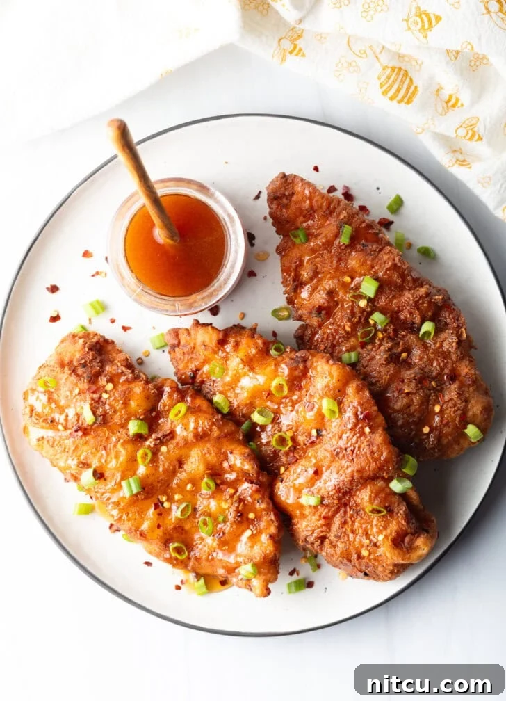 Sweet Heat Chicken 11 A delicious top-down view of three hot honey fried chicken pieces, generously garnished with green onions. A rustic wooden spoon rests in a jar of honey in the background.
