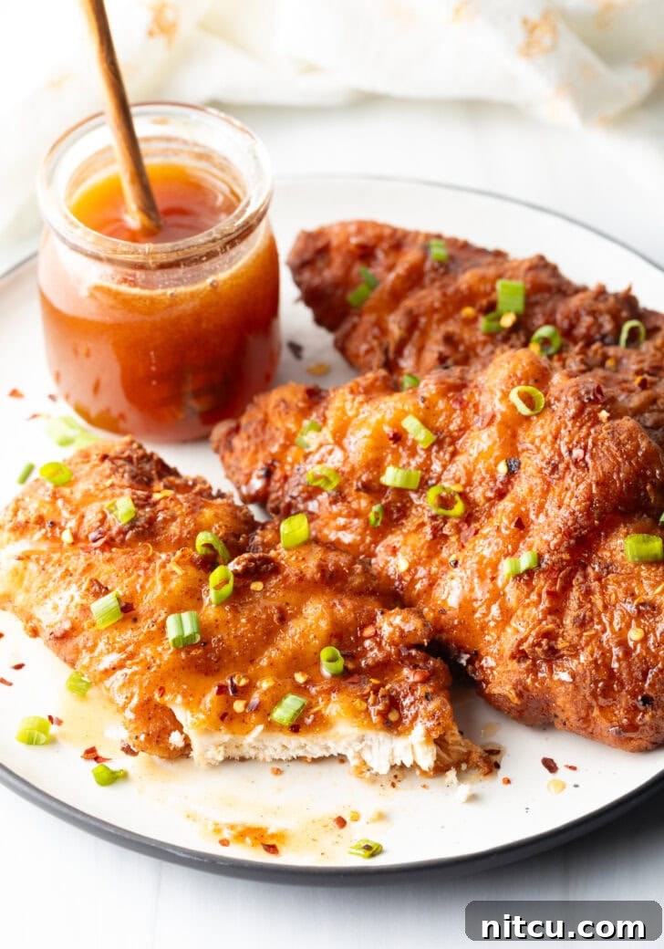 Sweet Heat Chicken 12 Three pieces of fried hot honey chicken, exquisitely plated and garnished with sliced green onions. A jar of honey sits in the background, and the front piece of chicken is cut open, revealing its tender, juicy interior.