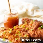 Sweet Heat Chicken 13 A close-up of three pieces of fried hot honey chicken, beautifully arranged on a plate with fresh green onions. A jar of honey is present in the background.