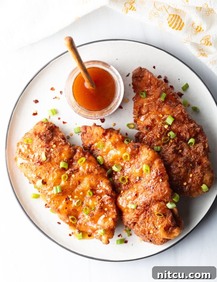 Sweet Heat Chicken 3 A top-down perspective of three glistening pieces of fried hot honey chicken, adorned with fresh green onions. A jar of honey and a wooden spoon are softly blurred in the background.