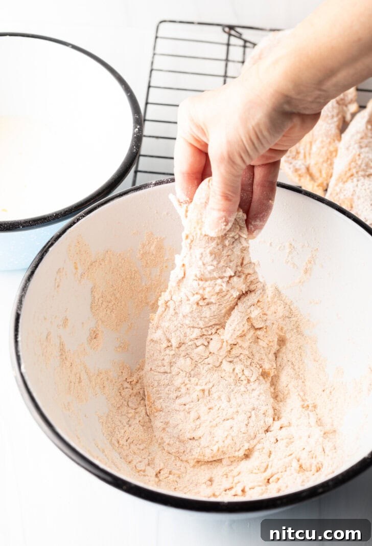 Sweet Heat Chicken 6 A hand gently coating a raw chicken breast in a bowl of white flour, demonstrating the initial dredging step for even coverage.