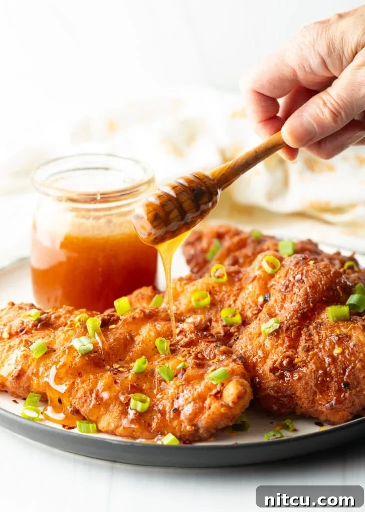 Sweet Heat Chicken 10 A mesmerizing shot of hot honey sauce being drizzled over three perfectly fried chicken pieces, garnished with green onions. A jar of honey is prominently featured in the background.