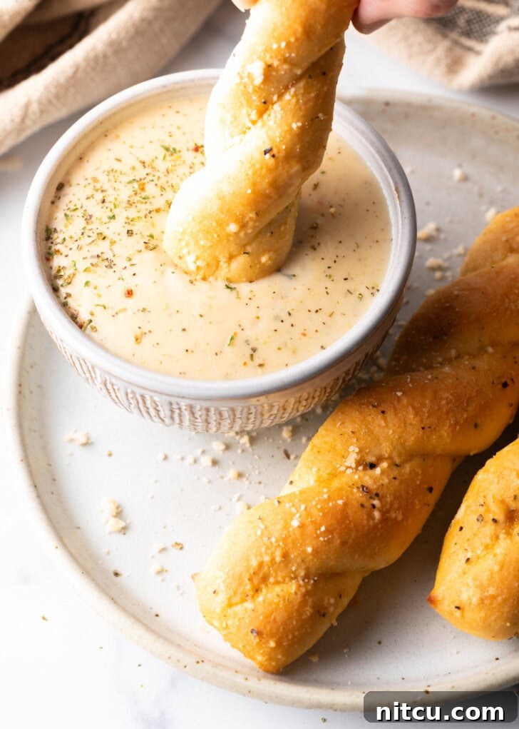 Top down view of a golden breadstick being dipped into a ramekin filled with rich, creamy garlic parmesan sauce, emphasizing its perfect dipping consistency.
