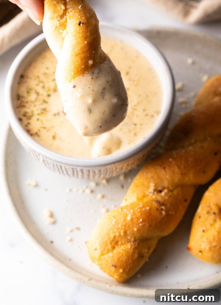Top down view of a golden breadstick being dipped into a ramekin filled with rich, creamy garlic parmesan sauce.