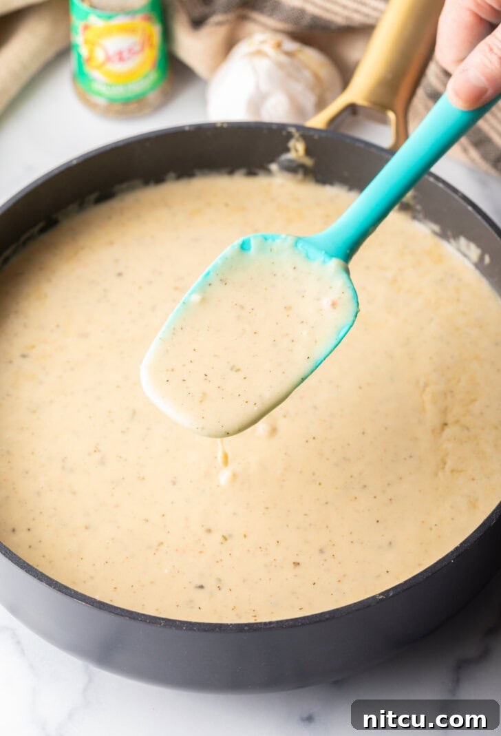 A blue spatula diligently stirring the creamy, bubbling garlic parmesan sauce in a skillet, ensuring a smooth, even consistency.