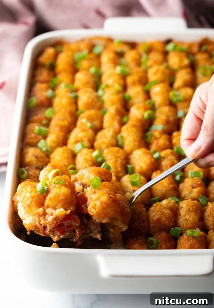 Chuckwagon Tater Tot Casserole 2 A fully baked Cowboy Tater Tot Casserole, showcasing rows of perfectly golden tater tots on top. A hand with a metal spoon scoops a generous serving from the dish.