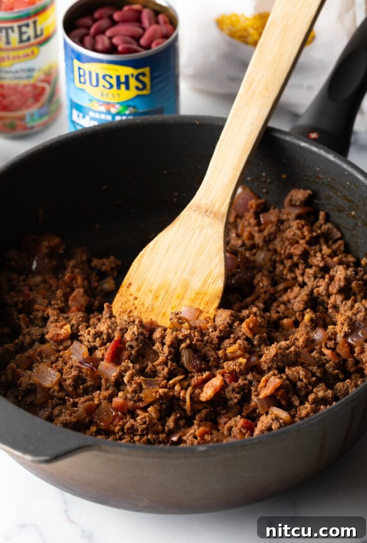 Chuckwagon Tater Tot Casserole 6 Deep black skillet with ground beef mixture, being stirred by a wooden spatula.