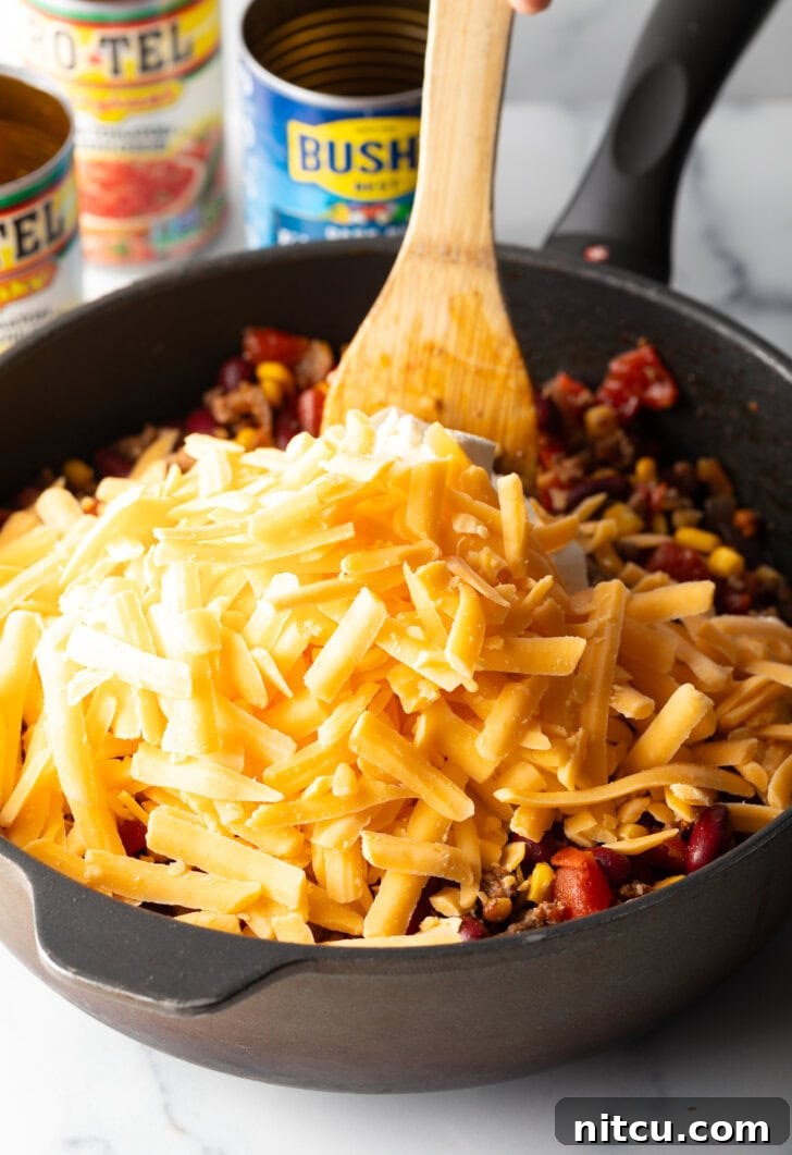Chuckwagon Tater Tot Casserole 8 Deep black skillet with ground beef mixture and shredded cheese, being stirred by a wooden spatula.