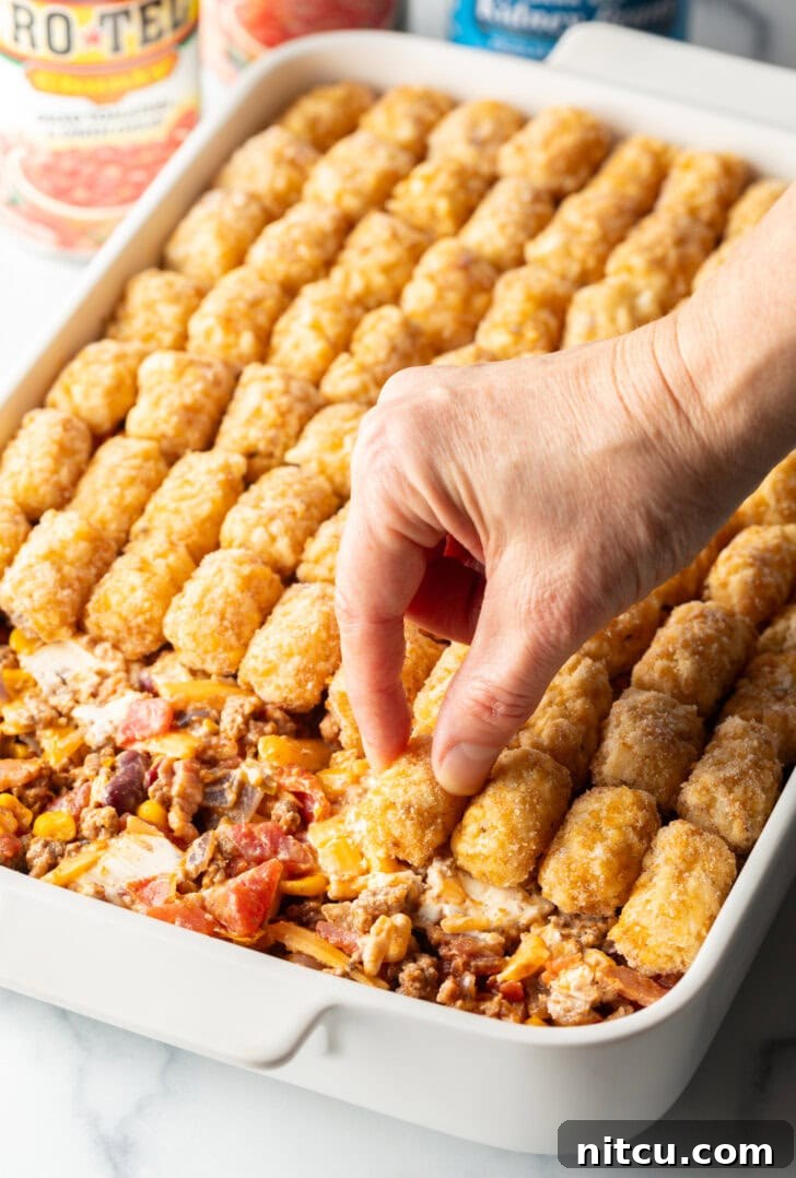 Chuckwagon Tater Tot Casserole 9 Hand adding frozen tater tots to the top of a baking dish casserole.