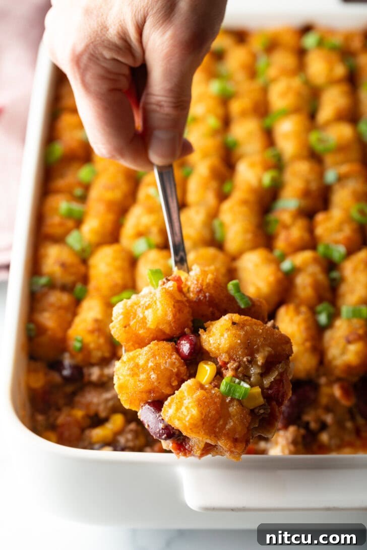 Chuckwagon Tater Tot Casserole 10 Full baked casserole with rows of tater tots on top. A hand with a metal spoon is scooping a serving.