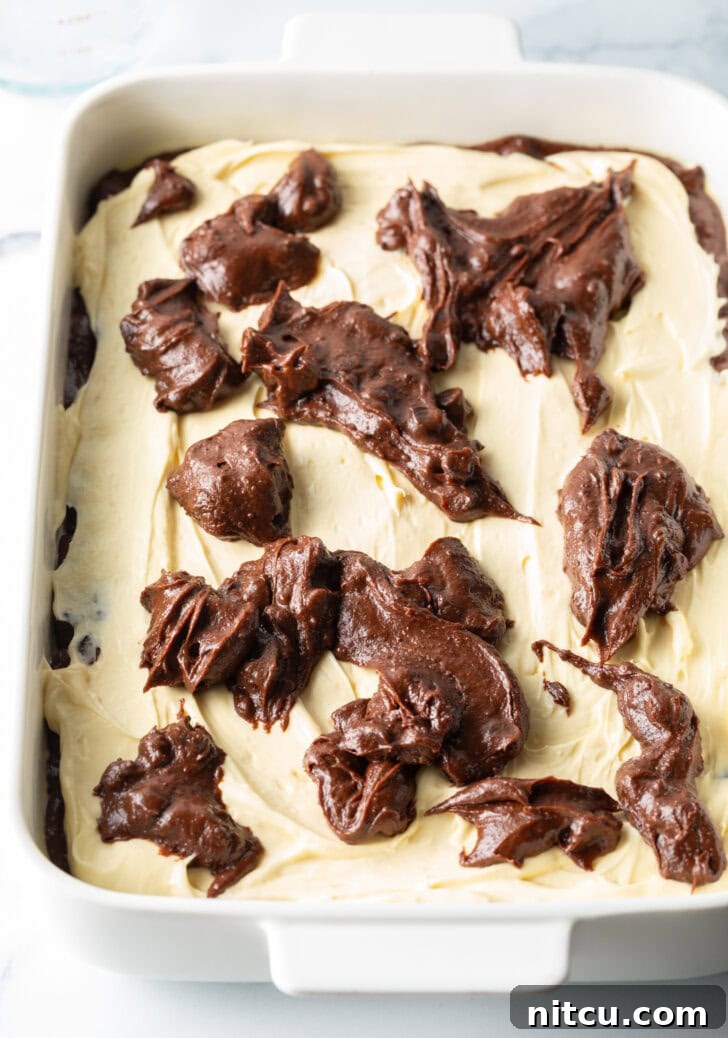 Decadent Brownie Cheesecake Bars 11 A top-down view of a white rectangular baking pan filled with layers of brownie batter, topped with cream cheese, and further dotted with dollops of more brownie batter, ready for swirling.