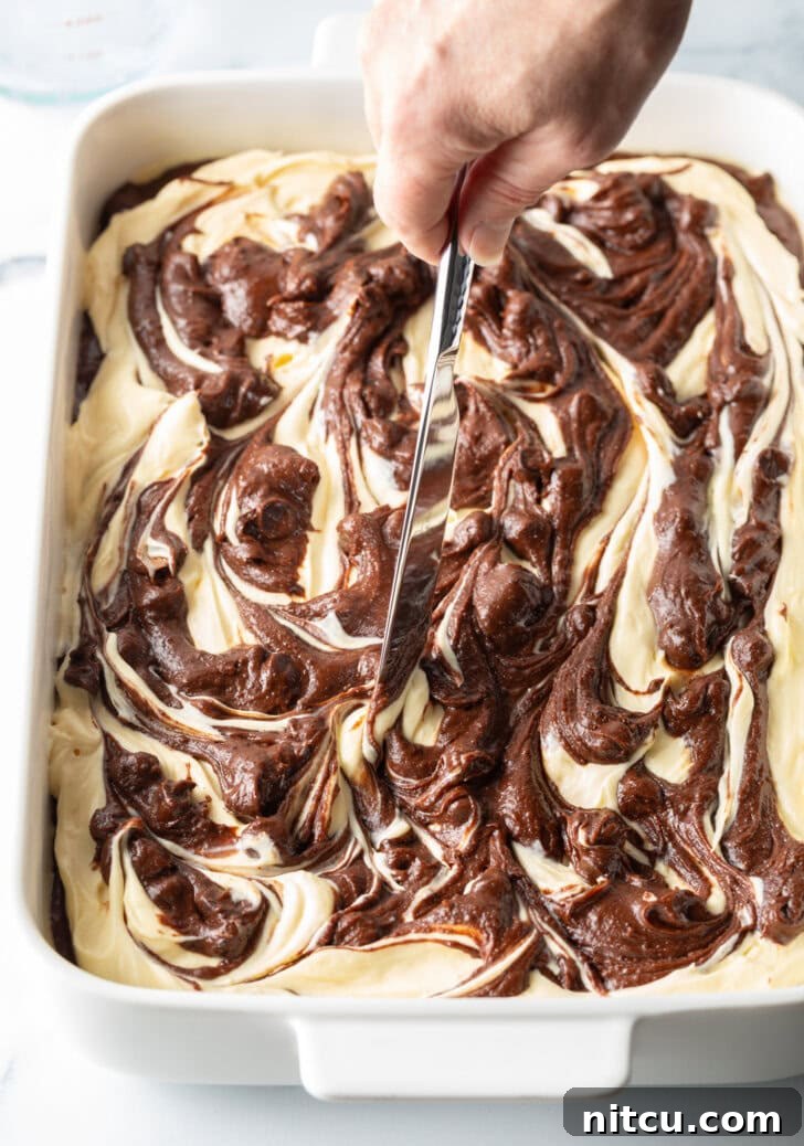 Decadent Brownie Cheesecake Bars 12 A hand uses a knife to artfully swirl the raw cheesecake and brownie batter together, creating a beautiful marbled pattern.
