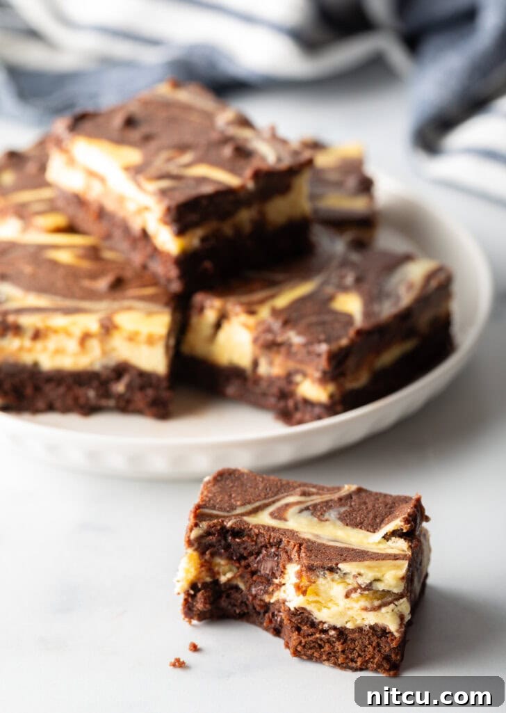Decadent Brownie Cheesecake Bars 16 A charming plate displays five cheesecake brownies, with one elegantly stacked, and a sixth brownie positioned closer to the camera for an inviting look.