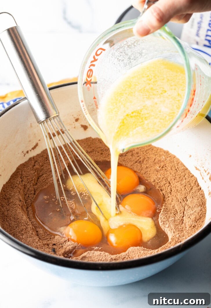 Decadent Brownie Cheesecake Bars 6 Melted butter is being poured from a glass measuring cup into a white bowl containing dry ingredients and cracked eggs, ready to be mixed into brownie batter.