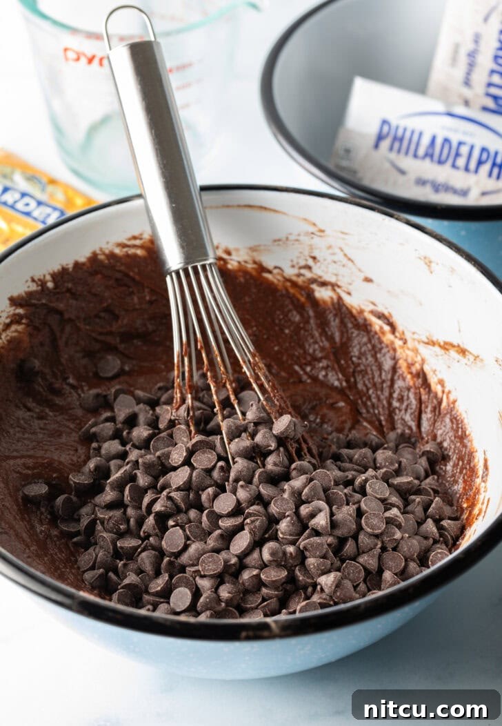 Decadent Brownie Cheesecake Bars 7 A whisk is gently incorporating chocolate chips into rich brownie batter in a pristine white mixing bowl.