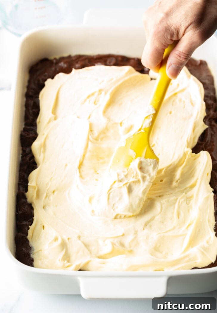 Decadent Brownie Cheesecake Bars 10 A hand uses a yellow spatula to gently spread a creamy cheesecake filling over the top of a layer of brownie batter in a baking pan.