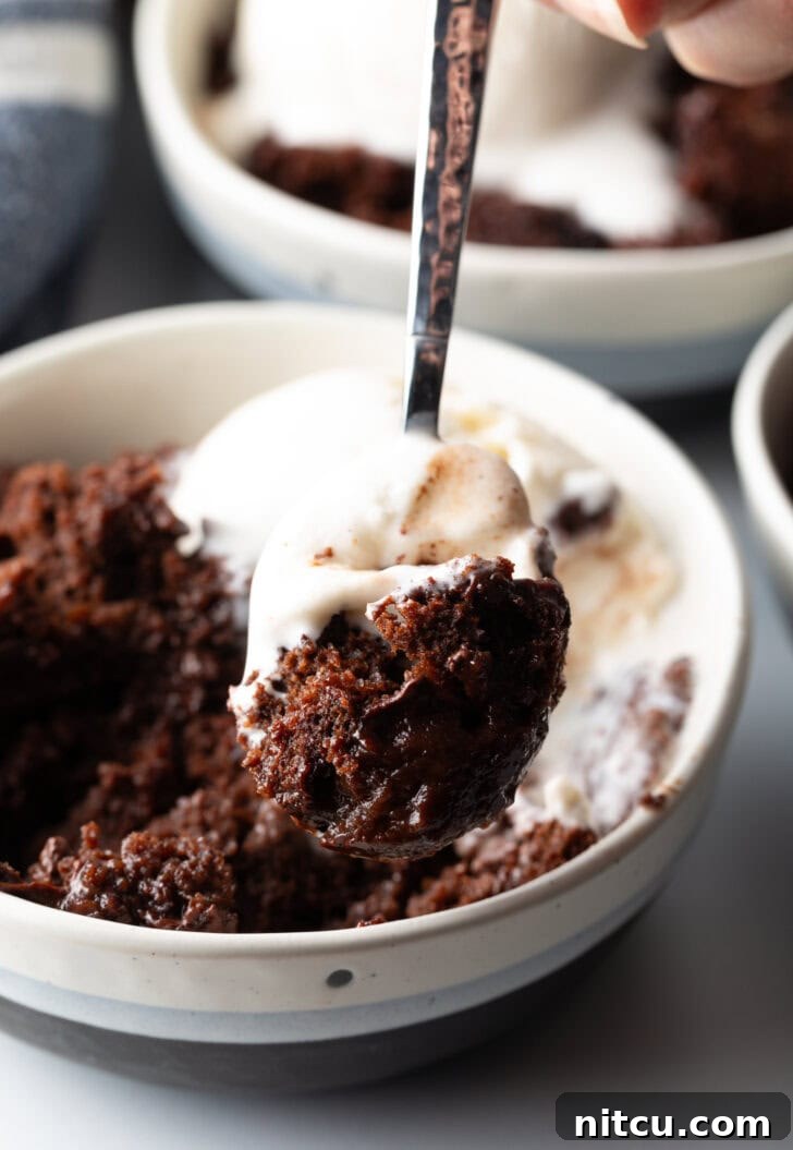 Decadent Chocolate Pudding Cake 13 White bowl with serving of chocolate lava cake and a scoop of vanilla ice cream. A hand with a metal spoon is taking a scoop from the bowl.