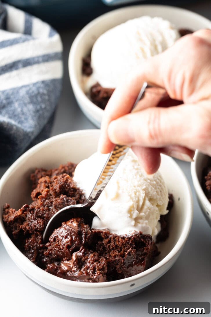 Decadent Chocolate Pudding Cake 4 White bowl with serving of chocolate lava cake and a scoop of vanilla ice cream. A hand with a metal spoon is taking a scoop from the bowl.