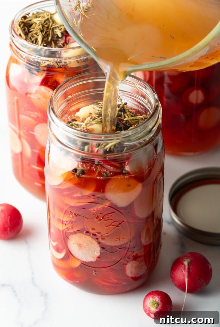 Hot pickling liquid being carefully poured from a glass measuring pitcher into a glass jar filled with radish slices and spices.
