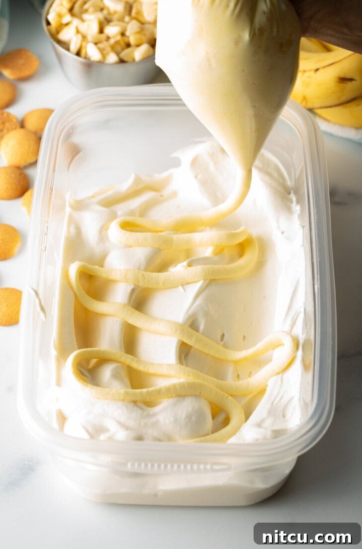Effortless Banana Pudding Ice Cream 7 A hand expertly squeezing rich banana pudding from a piping bag onto a layer of frozen ice cream within a rectangular freezer container, creating a beautiful swirl.