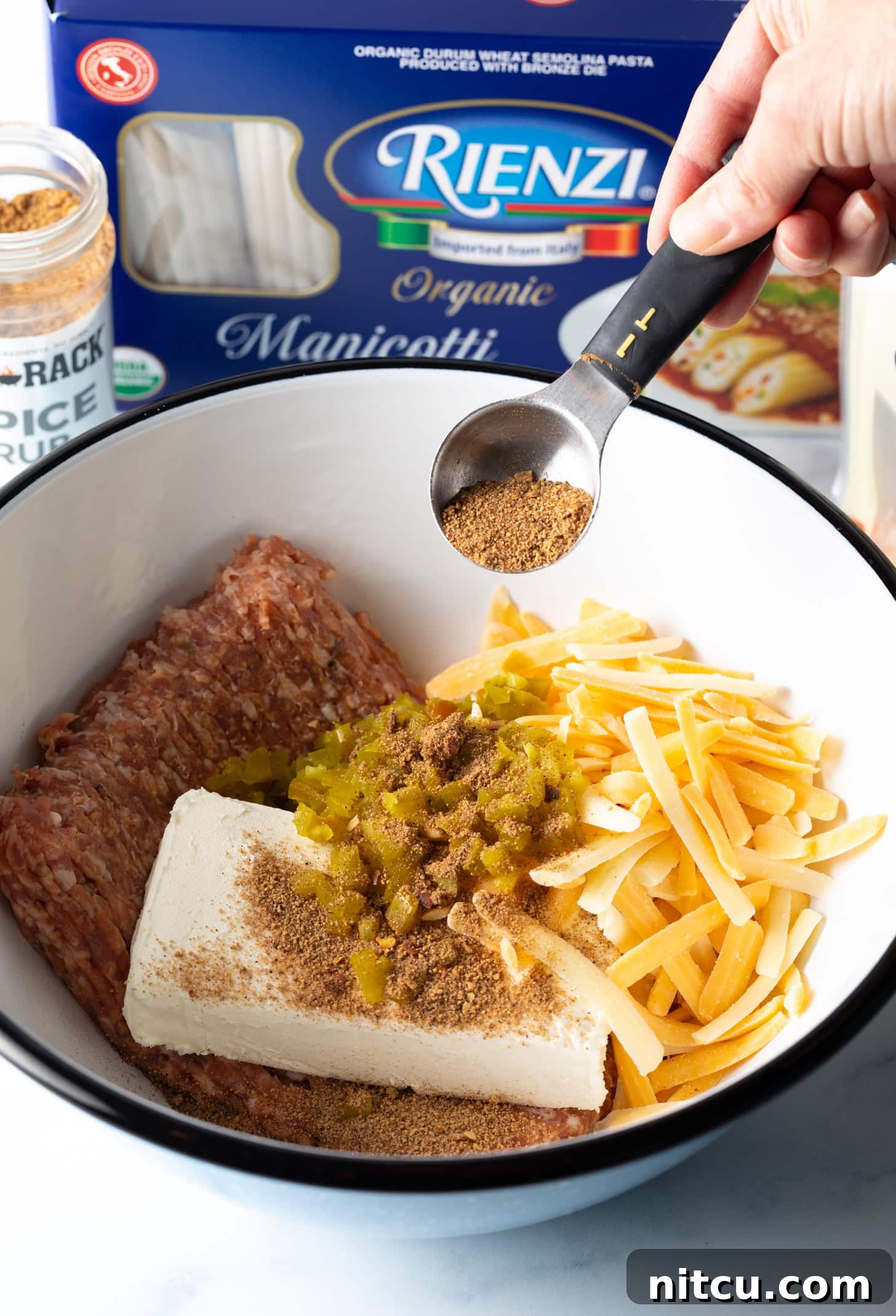 Smoked & Loaded Shells 6 Hand adding tablespoon of seasoning to a white bowl with raw ground beef, shredded cheddar cheese, block of cream cheese, and chopped peppers.