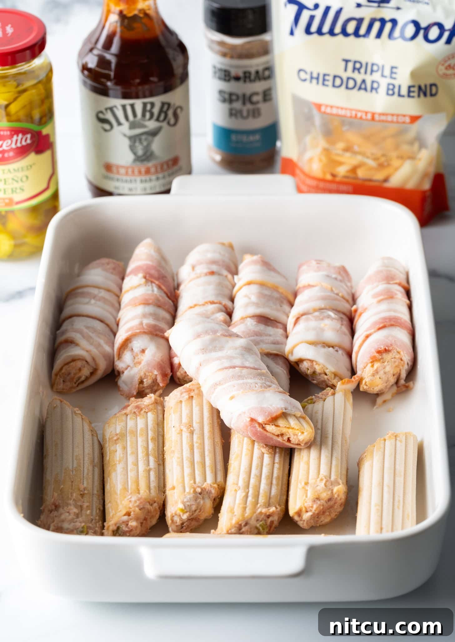 Smoked & Loaded Shells 7 White baking dish with two rows of meat and cheese stuffed manicotti. Half of the pasta is wrapped in pieces of raw bacon.