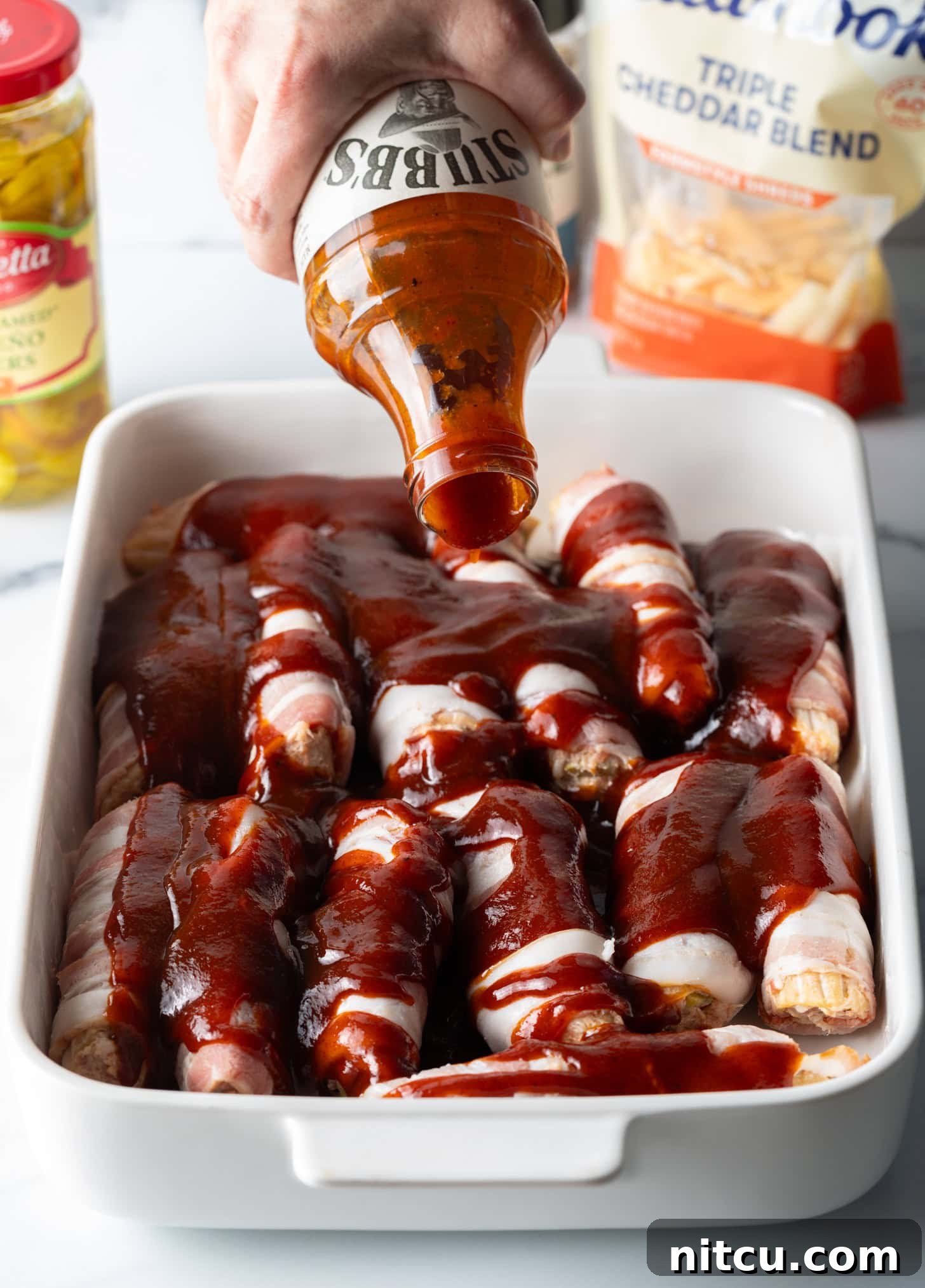 Smoked & Loaded Shells 8 Hand pouring barbecue sauce from a bottle on top of the bacon wrapped and stuffed shells layered in a white baking dish.