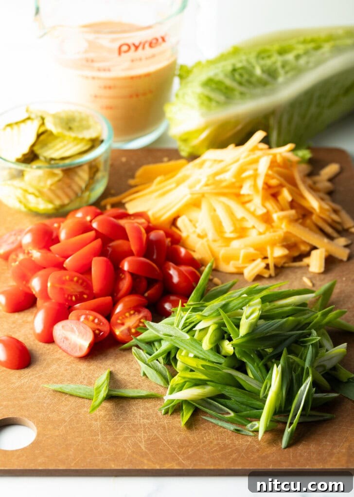 Big Mac Burger Bowl 5 Recipe ingredients on a cutting board: Neat piles of bright shredded cheese, halved red cherry tomatoes, vibrant sliced green onions, and a small glass brimming with crisp pickle slices, highlighting fresh preparation.