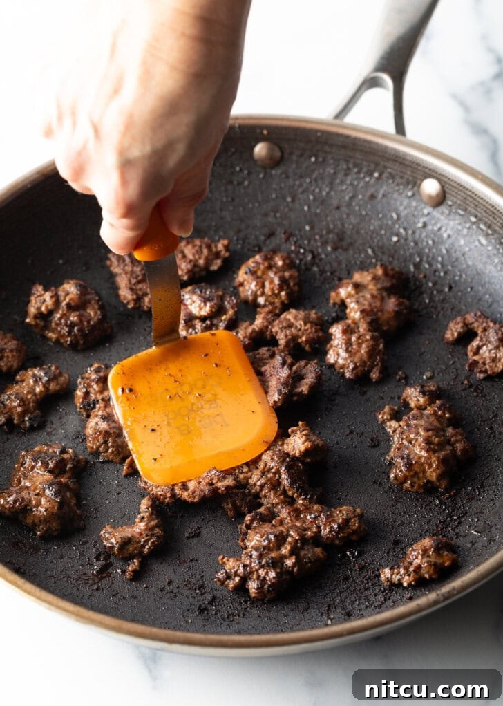 Big Mac Burger Bowl 6 A hand with a spatula expertly cooking mini burger patties in a hot skillet, ensuring a perfect sear and delicious browned crumbles for the salad.