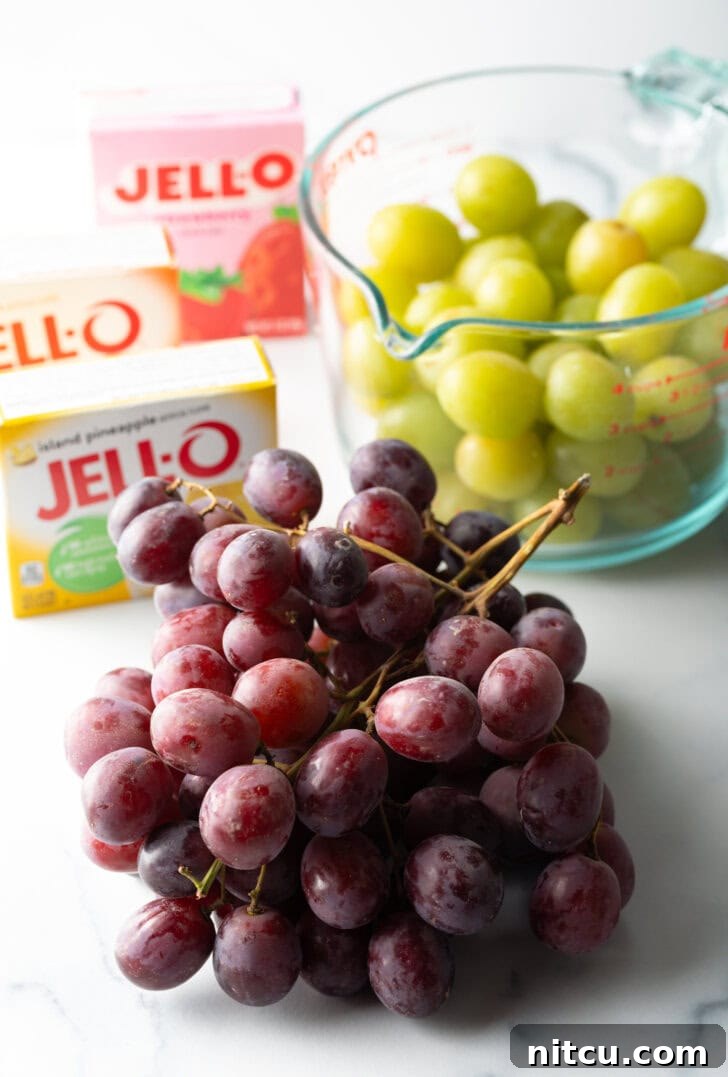 Sweet Grape Gems 6 Recipe ingredients: a bunch of red grapes in the front view, in the background is a large glass measuring cup with green grapes. 3 boxes of Jello are to the side.