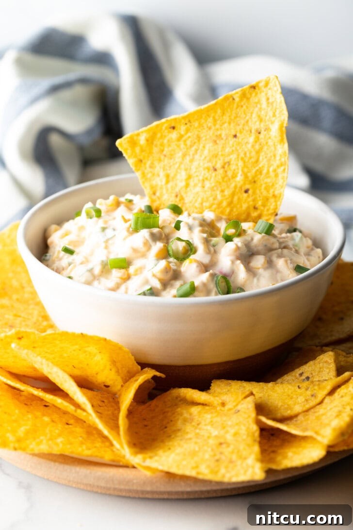 Creamy Street Corn Dip 8 A tortilla chip loaded with creamy Mexican corn dip being scooped from a pristine white bowl, showcasing its rich texture.