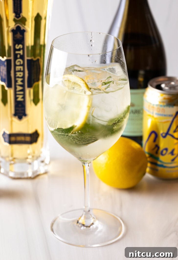 St-Germain Garden Spritz 2 Wine glass with alcohol, mint leaves, and lemon slices. In the background of the hugo spritz are a whole lemon and bottles of prosecco, st. germain liquor, and a can of la croix soda.