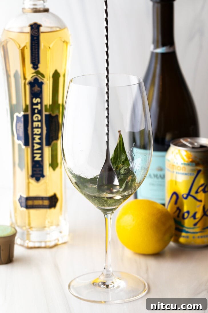 St-Germain Garden Spritz 5 Wine glass with mint leaves being muddled with a metal spoon.