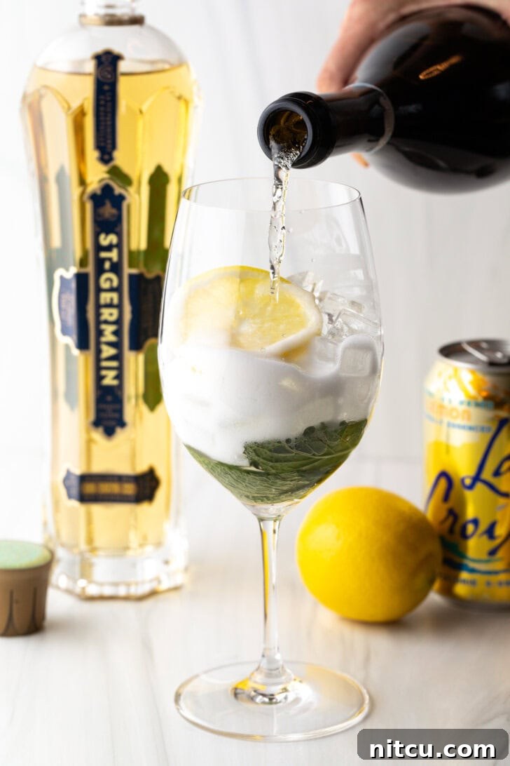 St-Germain Garden Spritz 7 Wine glass with alcohol, mint leaves, and lemon slices. In the background are a whole lemon, st. germain liquor, and a can of la croix soda. A hand is adding prosecco from a bottle.