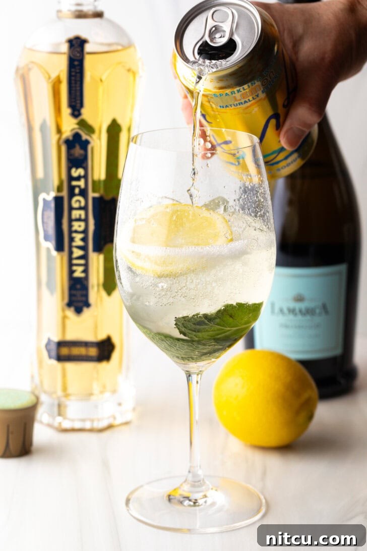 St-Germain Garden Spritz 8 Wine glass with alcohol, mint leaves, and lemon slices. In the background are a whole lemon and bottles of champagne, st. germain liquor. A hand is adding soda from a can of la croix soda.