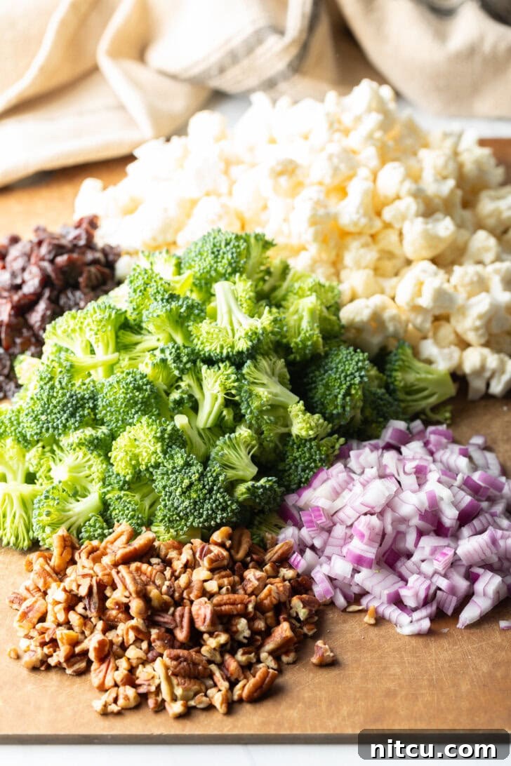 Vibrant Broccoli Cauliflower Salad 5 Cutting board showcasing neatly piled fresh broccoli florets, cauliflower florets, finely chopped red onions, crunchy pecans, and bright dried cranberries, ready for salad assembly.