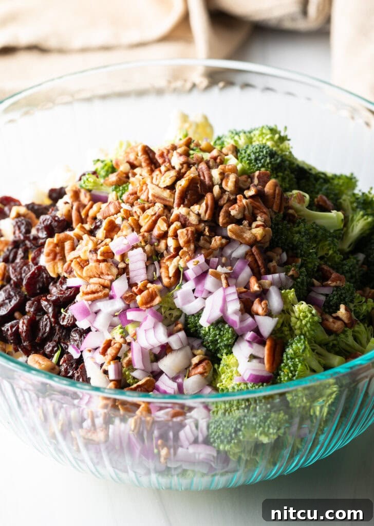 Vibrant Broccoli Cauliflower Salad 6 A large glass bowl filled with fresh broccoli florets, cauliflower florets, chopped red onions, pecans, and cranberries, awaiting the dressing.