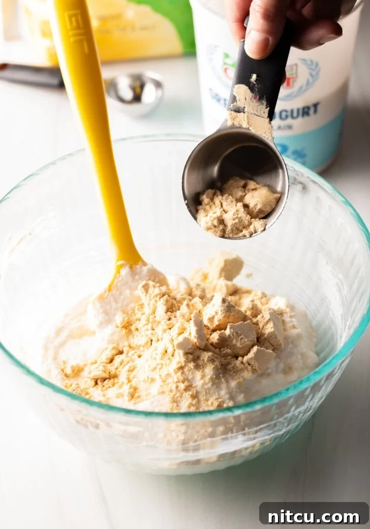 High-Protein Yogurt Pudding 5 Adding protein powder from a metal measuring cup into a glass bowl with a yellow spatula.