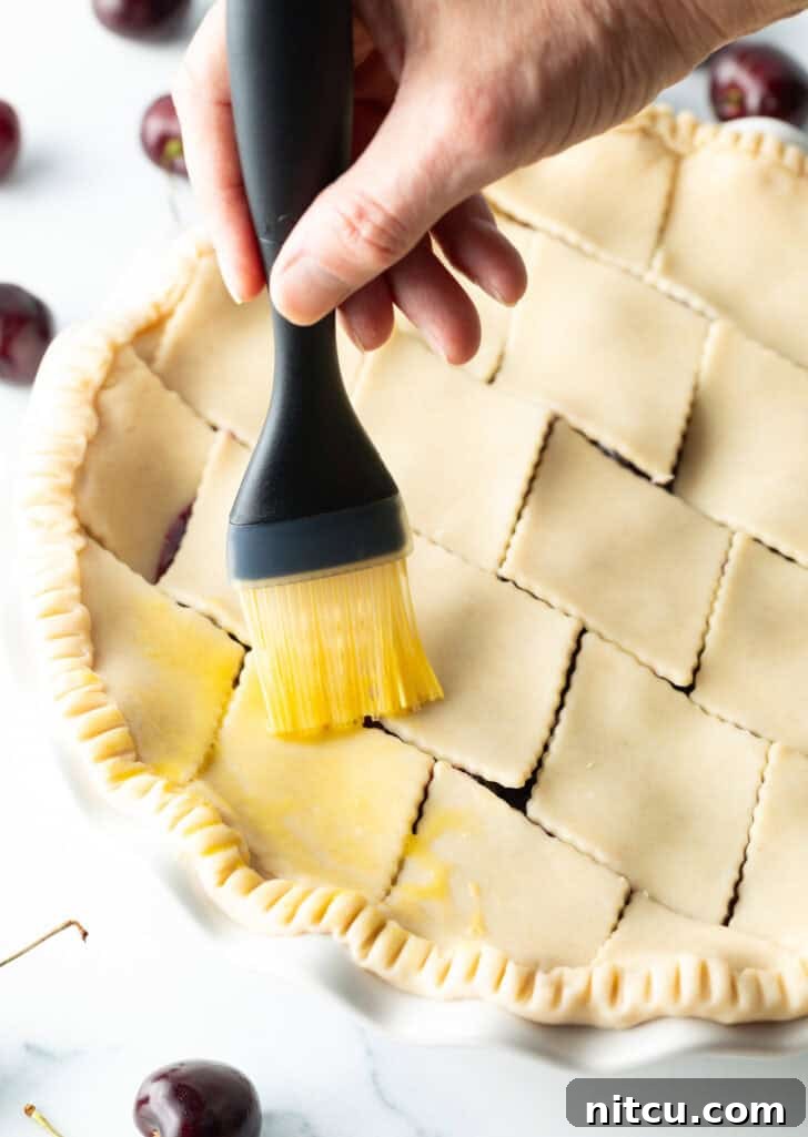 Sweet Cherry Pie Delight 10 A hand gently brushes an egg wash over the intricate pattern of an unbaked pie's top crust, preparing it for a golden, glossy finish.
